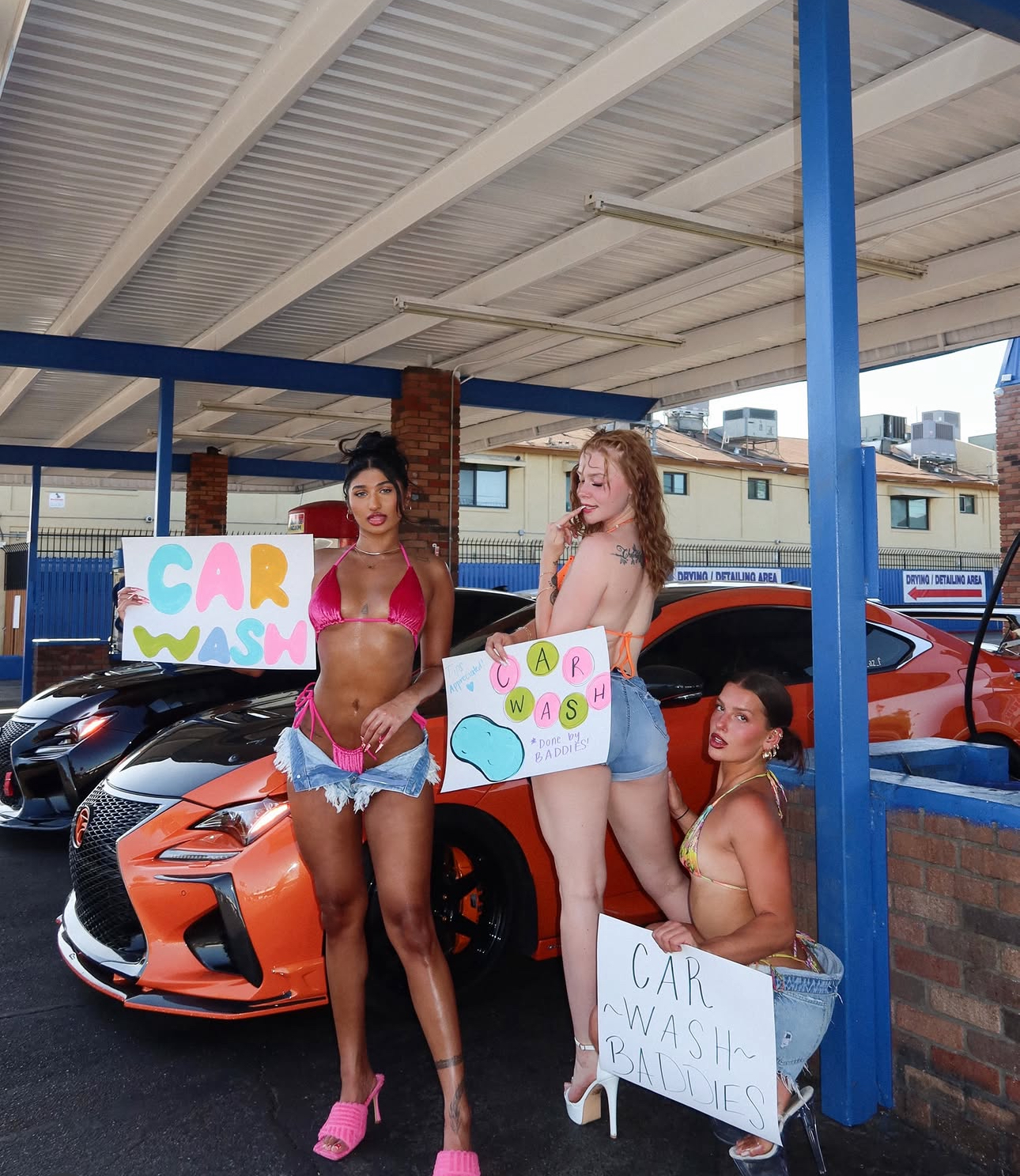 Three Arys Grrls members in bikinis and denim shorts holding hand-painted car wash signs beside a bright orange sports car